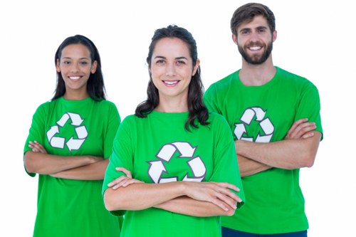 Three individuals wearing bright green T-shirts with a white recycling symbol printed on the front are standing outdoors on a paved surface, likely in a residential or urban area. The woman in the centre has dark, wavy hair pulled back and is smiling directly at the camera, with her arms crossed. To her left, a woman with long dark hair, also smiling, has her arms crossed and is positioned to partly face the camera. To her right, a man with a beard and short dark hair, also smiling, stands with his arms crossed, facing forward. The background is a plain, light-colored wall, suggesting an urban environment related to waste management or recycling activities in the vicinity of Amersham or nearby areas. Their attire and confident stance imply they are part of a community or waste management team involved in rubbish removal or recycling efforts, potentially representing a local initiative by House Clearance Amersham, which specialises in rubbish and home clearance services.