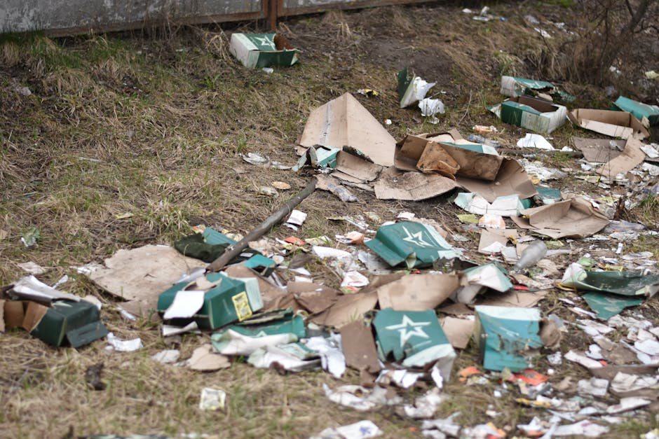 The image shows an outdoor area adjacent to a fence, where discarded cardboard boxes and packaging materials are scattered across the grassy ground. The boxes are mostly green with white star logos, i