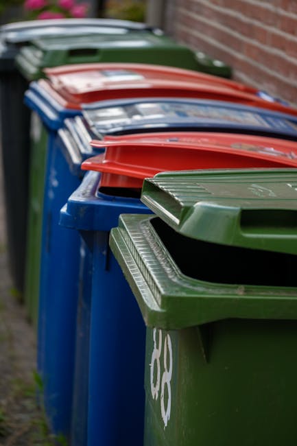 A row of several large wheelie bins lined up outdoors against a brick wall. The bins are made of plastic with textured surface patterns on their lids. The bins are arranged in a sequence from foregrou