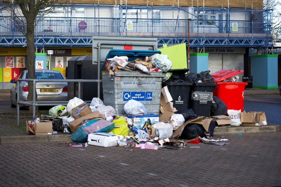 A pile of waste and rubbish-scattered debris lies on a gravel surface next to a low stone wall, with several black plastic rubbish bags, a yellow plastic storage container, and an old, dirty car tyre 